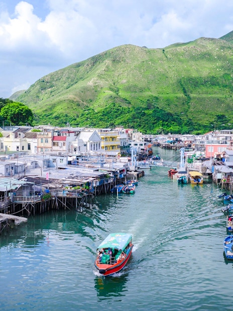 Boat navigating through Tai O fishing village with stilt houses and green hills in the background.