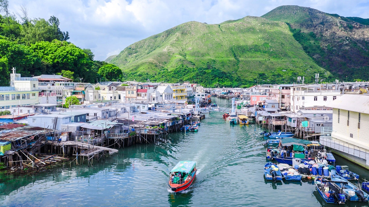 Boat navigating through Tai O fishing village with stilt houses and green hills in the background.