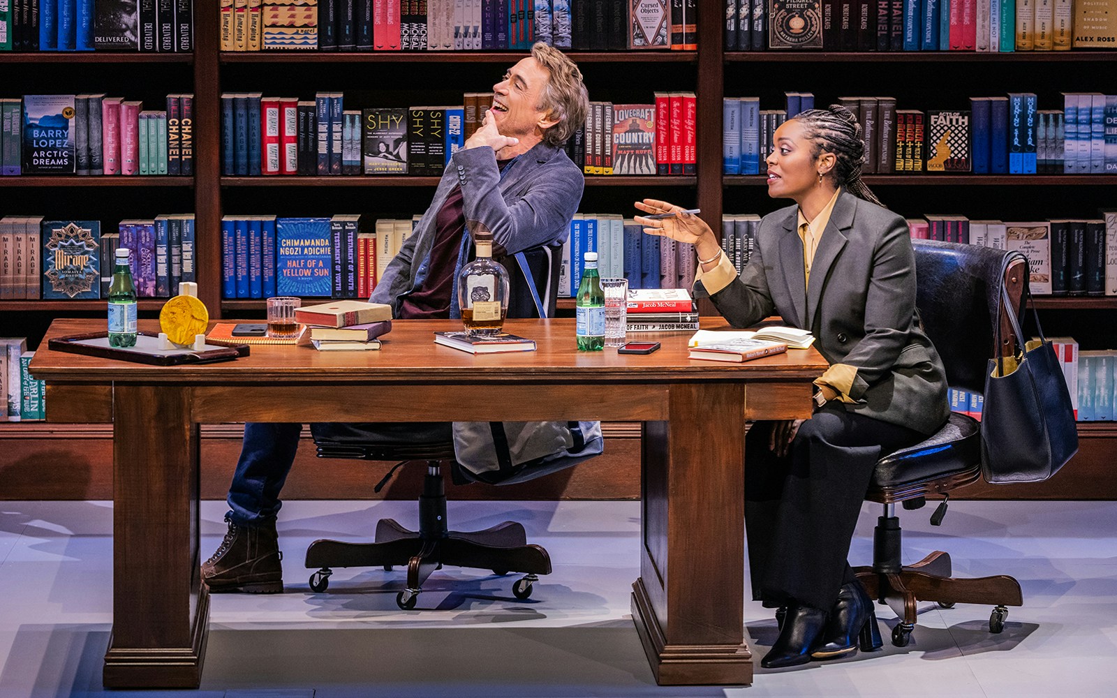 Two people sitting at a desk in a library, surrounded by books, engaged in conversation.