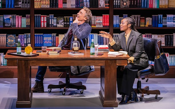 Two people sitting at a desk in a library, surrounded by books, engaged in conversation.