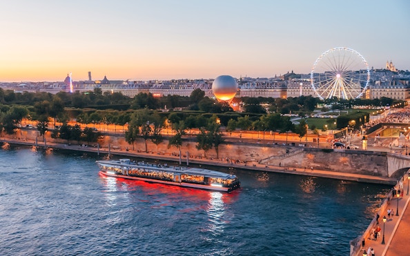 Seine River cruise boat passing by Paris landmarks at sunset with Ferris wheel in view.