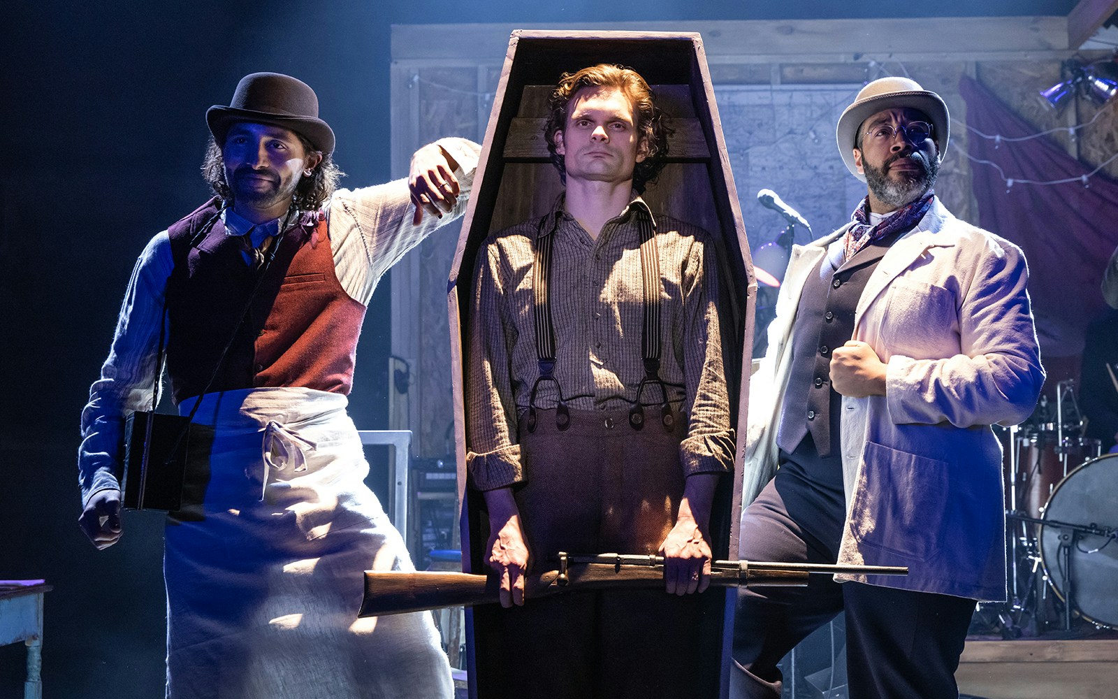 Three men in period costumes, one in a coffin holding a rifle, on stage during a "Dead Outlaw" performance.