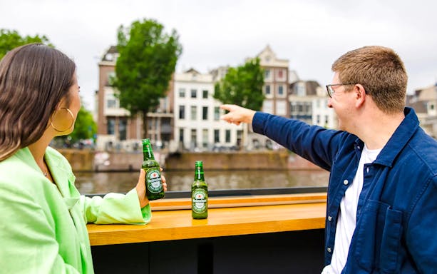 Tourists enjoying Heineken Experience flagship cruise on Amsterdam canal with cityscape views.