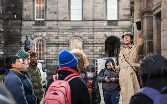 Tour guide in trench coat leading a group in Edinburgh, highlighting historical sites.