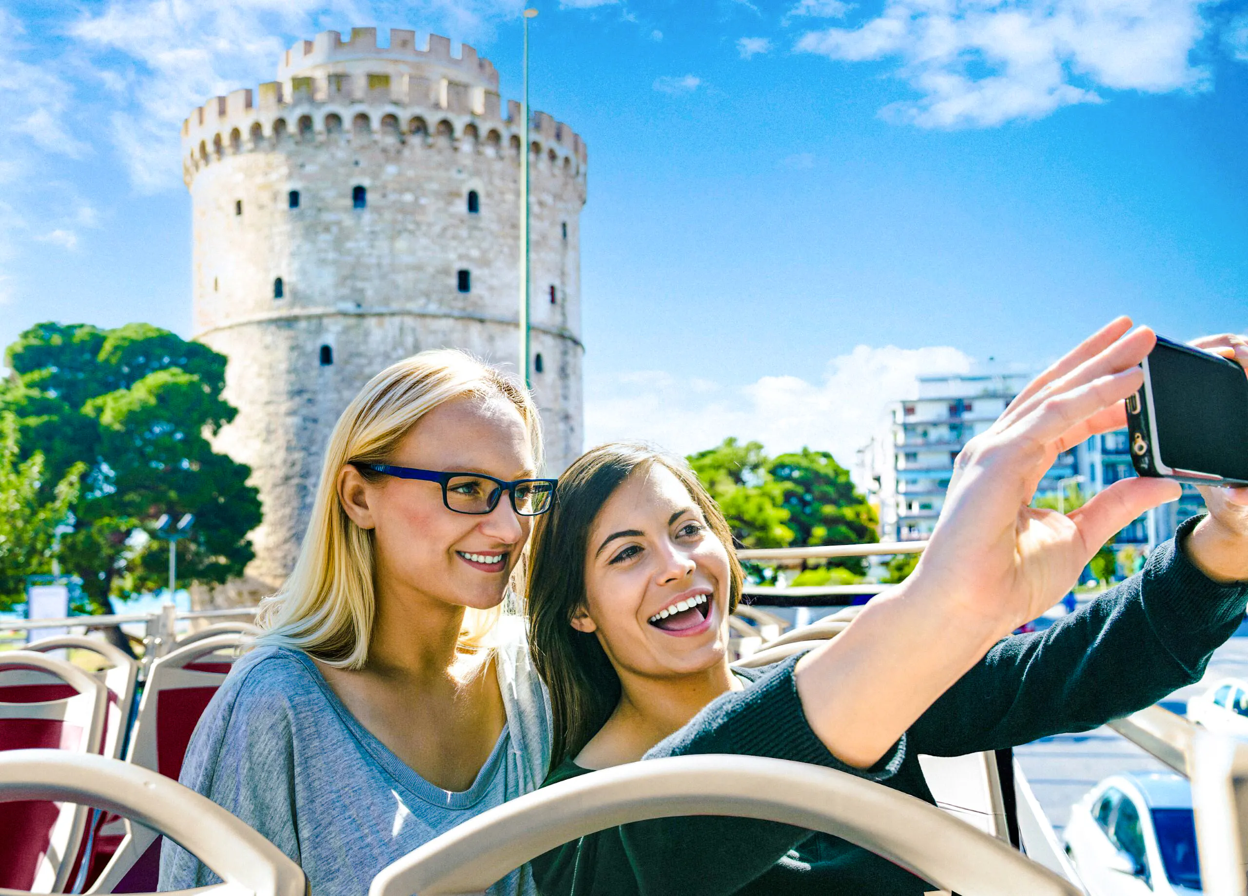 Two people taking a selfie on a bus with Thessaloniki's White Tower in the background.