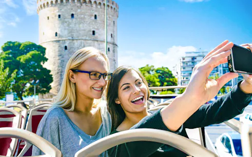 Two people taking a selfie on a bus with Thessaloniki's White Tower in the background.