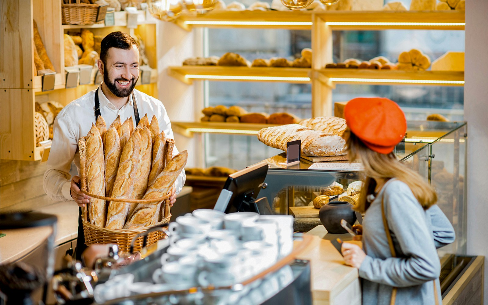 Baker holding basket of French bread in a bakery with customer at counter.