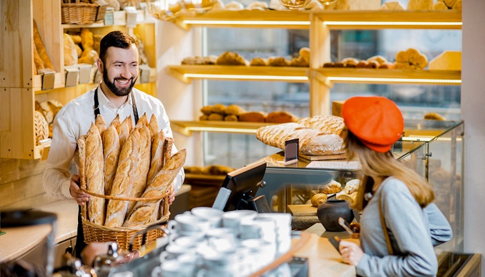 Baker holding basket of French bread in a bakery with customer at counter.