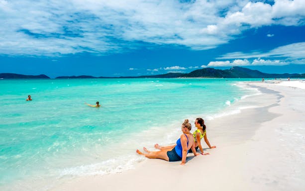 Tourists relaxing on Whitehaven Beach with turquoise water and distant hills.