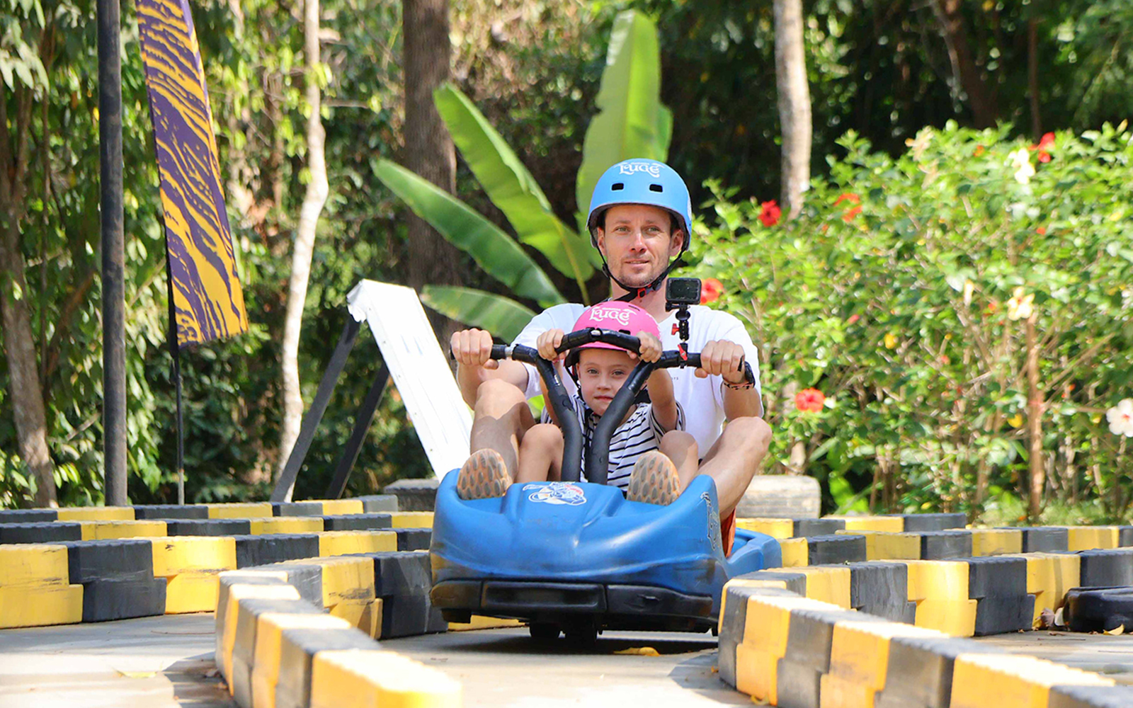 Father and child riding a blue go-kart at Hanuman World, surrounded by lush greenery.