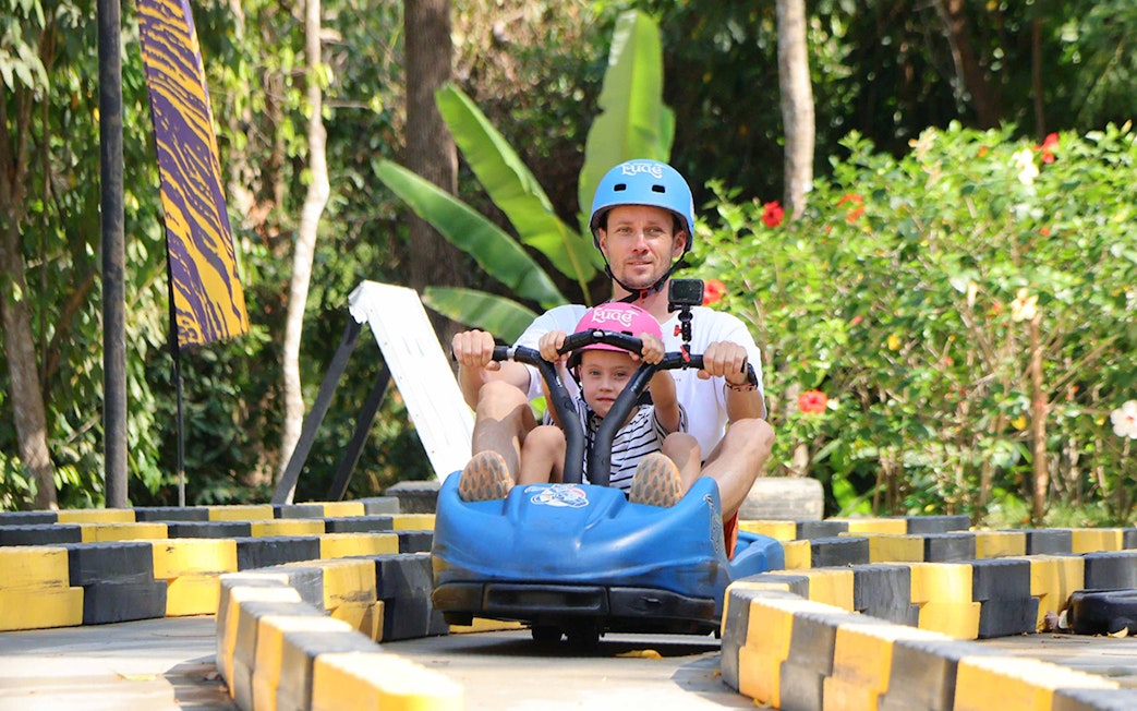 Father and child riding a blue go-kart at Hanuman World, surrounded by lush greenery.