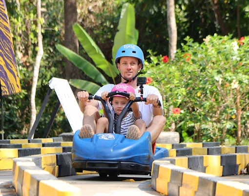 Father and child riding a blue go-kart at Hanuman World, surrounded by lush greenery.