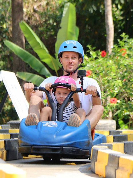 Father and child riding a blue go-kart at Hanuman World, surrounded by lush greenery.
