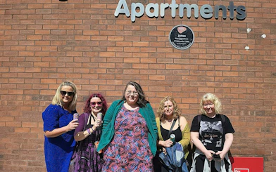 Group of people standing by a brick wall with a plaque in Manchester, related to 80s and 90s music tour.