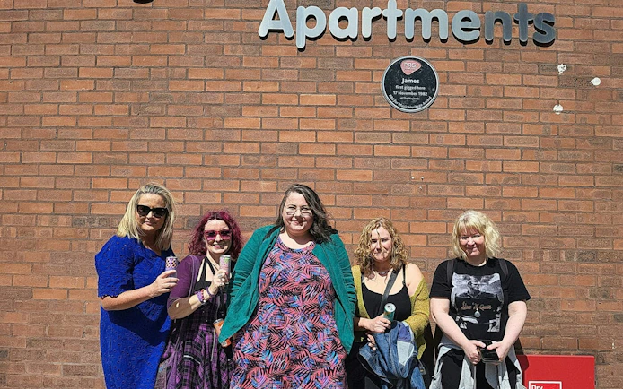 Group of people standing by a brick wall with a plaque in Manchester, related to 80s and 90s music tour.