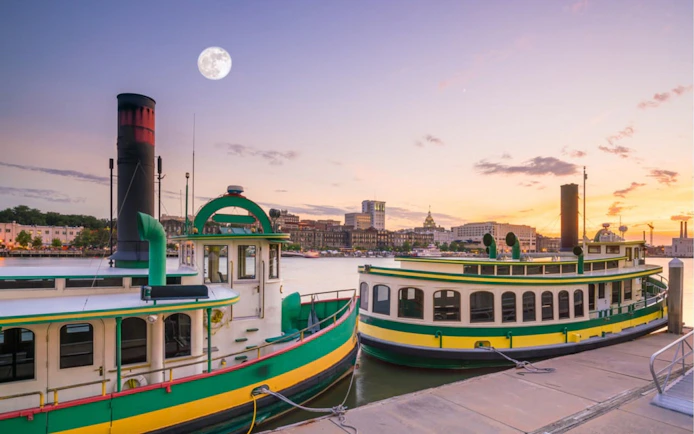 Savannah riverboats docked at sunset with city skyline in the background.
