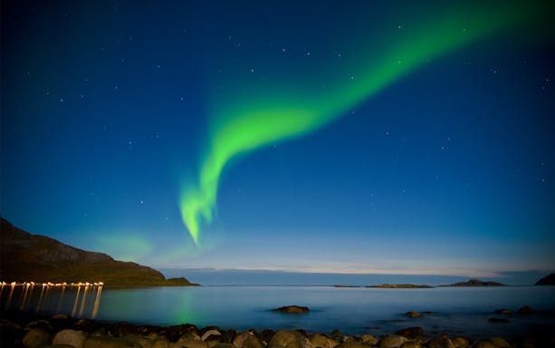 Northern Lights over calm sea viewed from cruise, rocky shoreline in foreground.