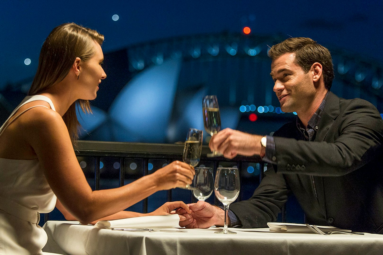 Couple toasting with champagne at a table overlooking Sydney Opera House at night.