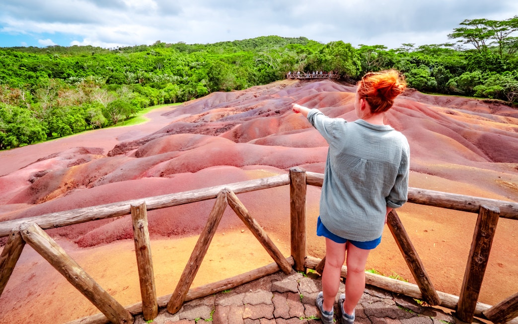 Visitor pointing at Chamarel 7 Coloured Earth in Mauritius with lush greenery in the background.