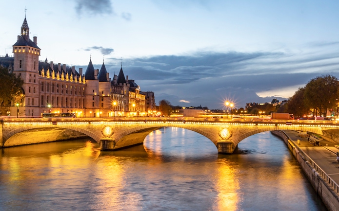 Conciergerie Paris illuminated at night with Seine River and bridge in view.