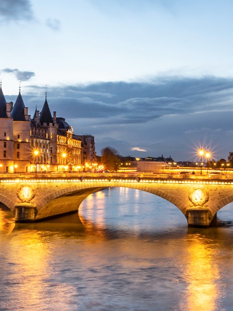 Conciergerie Paris illuminated at night with Seine River and bridge in view.