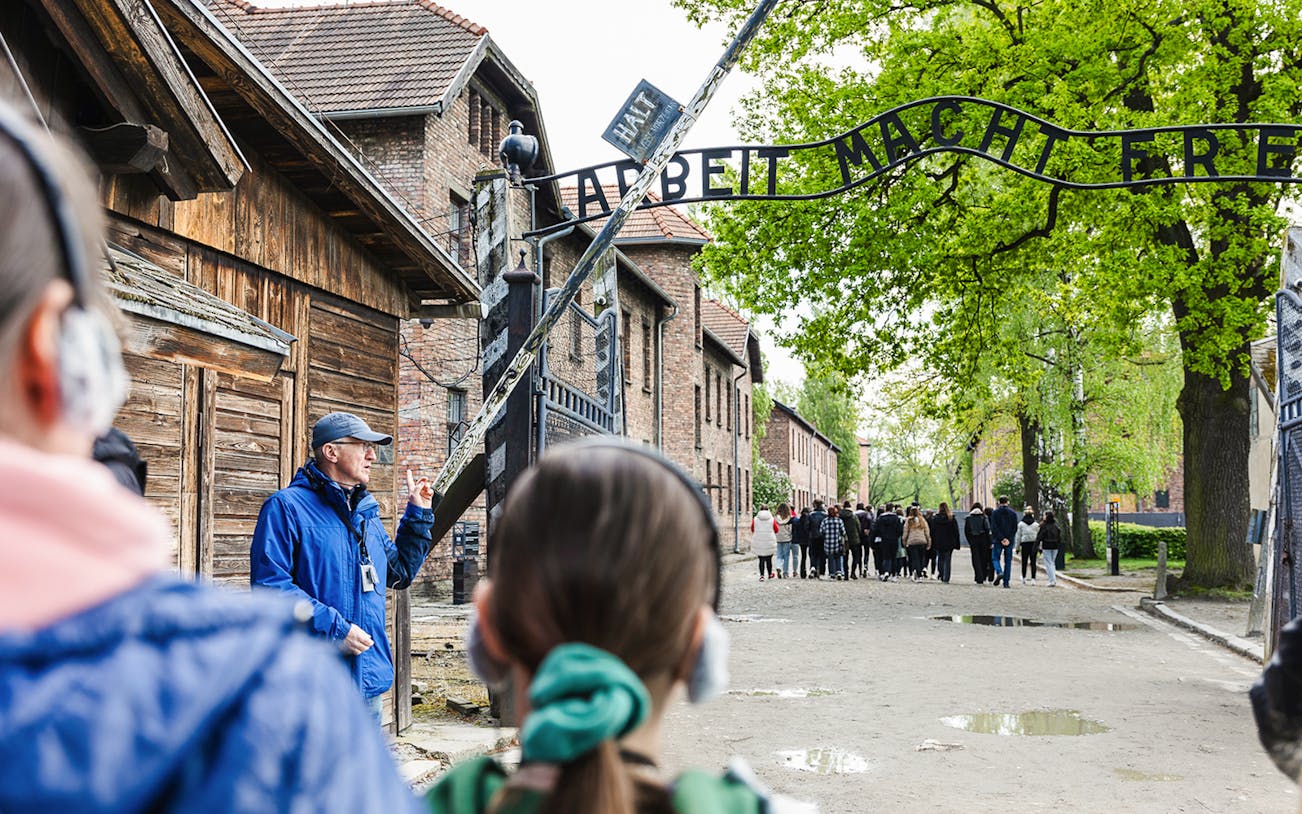 Guide leading a tour at the Auschwitz Birkenau entrance with visitors listening attentively.