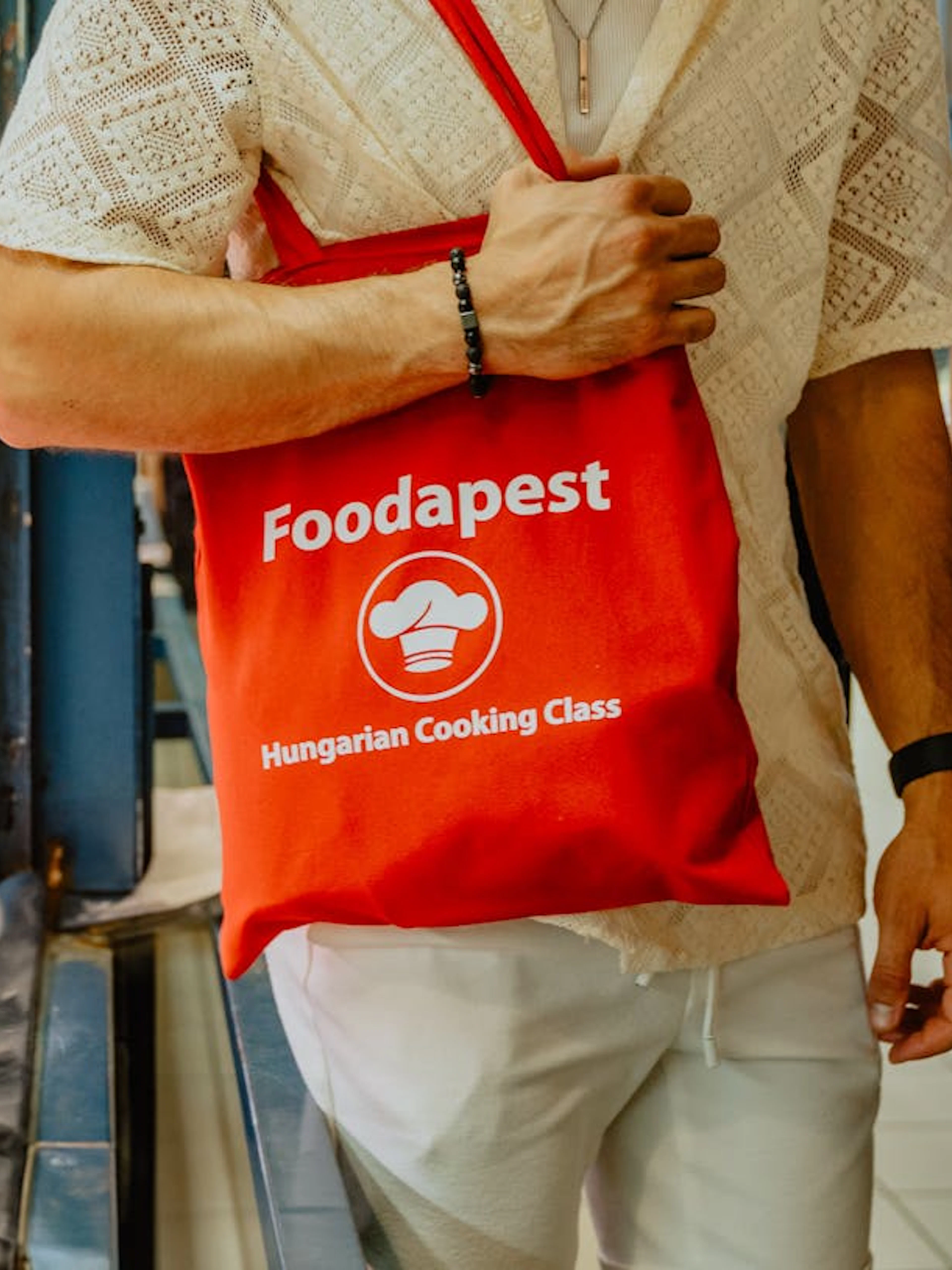 Man holding a "Foodapest Hungarian Cooking Class" bag in Budapest market.
