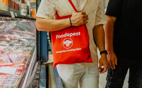 Man holding a "Foodapest Hungarian Cooking Class" bag in Budapest market.
