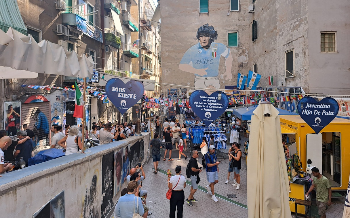 Street scene in Naples with a mural of a famous footballer, tourists exploring the area.