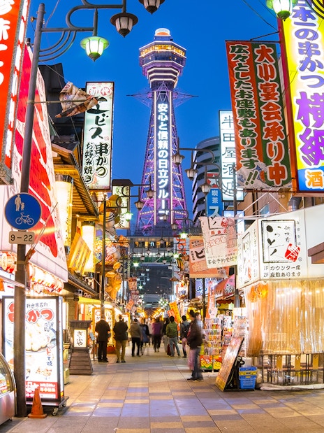 Bustling Osaka street with Tsutenkaku Tower, vibrant signs, and people exploring shops.