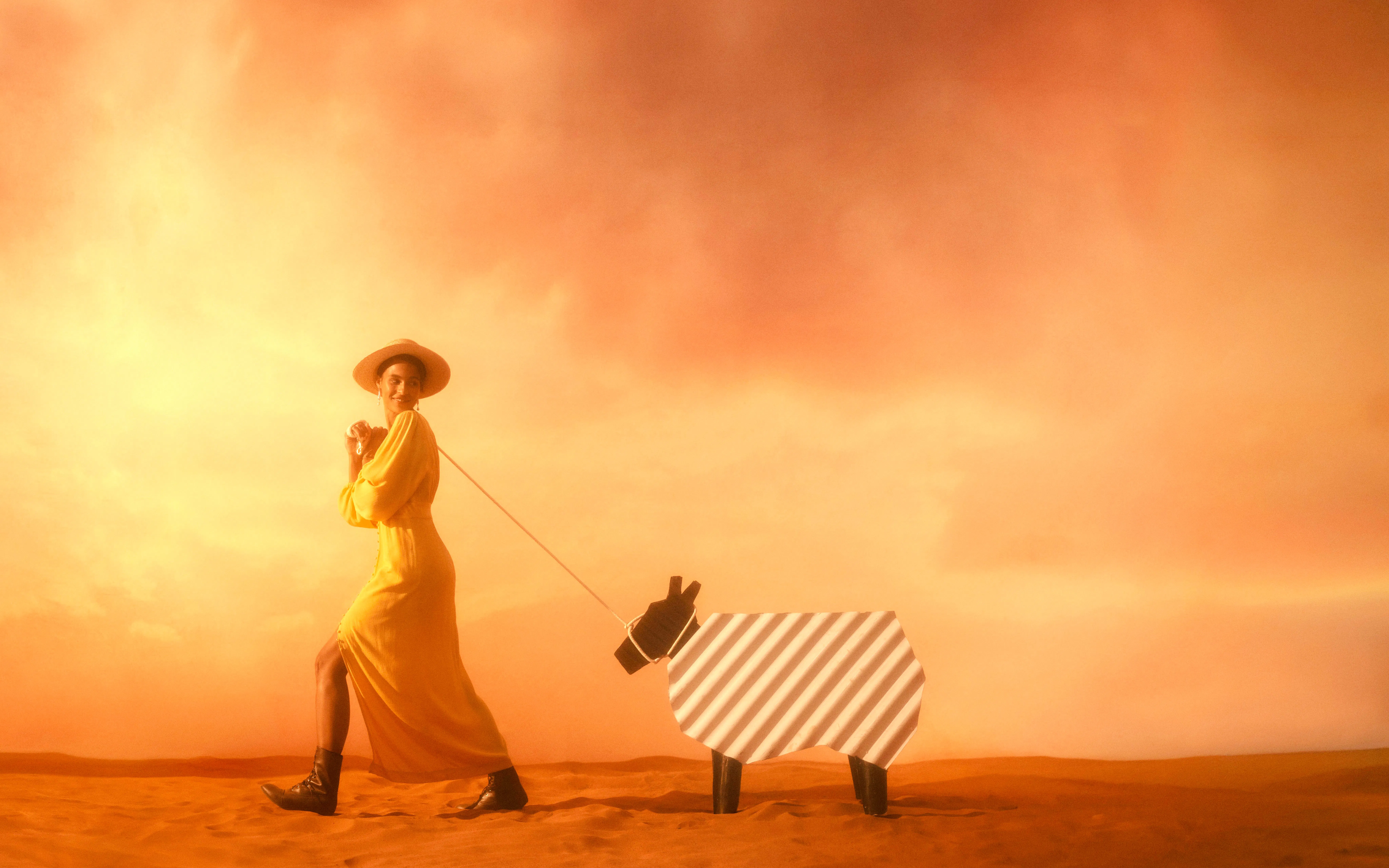 Person in a yellow dress walking a striped animal figure in a desert setting.