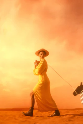 Person in a yellow dress walking a striped animal figure in a desert setting.