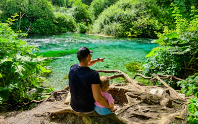 Father and toddler sitting by Blue Eye springs, surrounded by lush greenery.