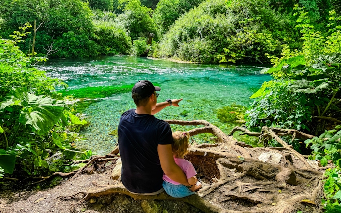 Father and toddler sitting by Blue Eye springs, surrounded by lush greenery.