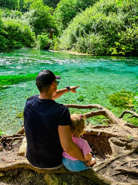 Father and toddler sitting by Blue Eye springs, surrounded by lush greenery.