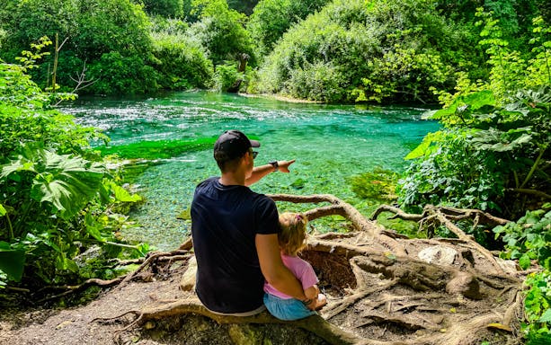 Father and toddler sitting by Blue Eye springs, surrounded by lush greenery.