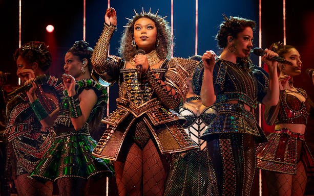 Performers in elaborate costumes singing on stage during SIX: The Musical.