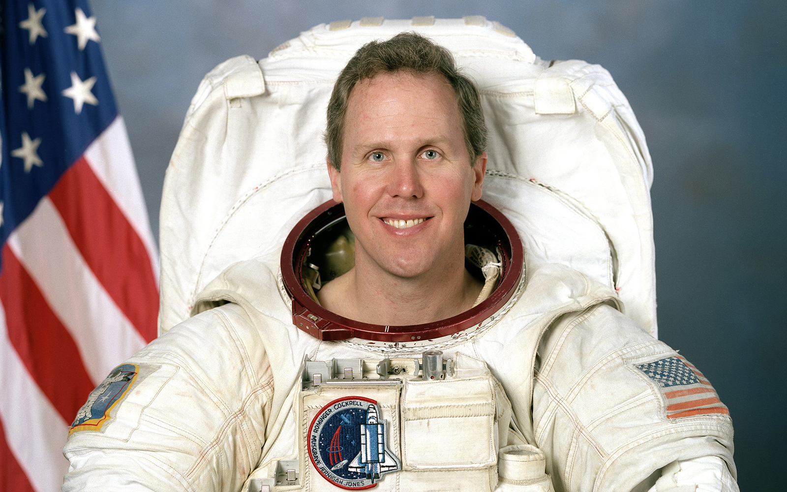 Astronaut Tom Jones in space suit with helmet, Earth visible in background.