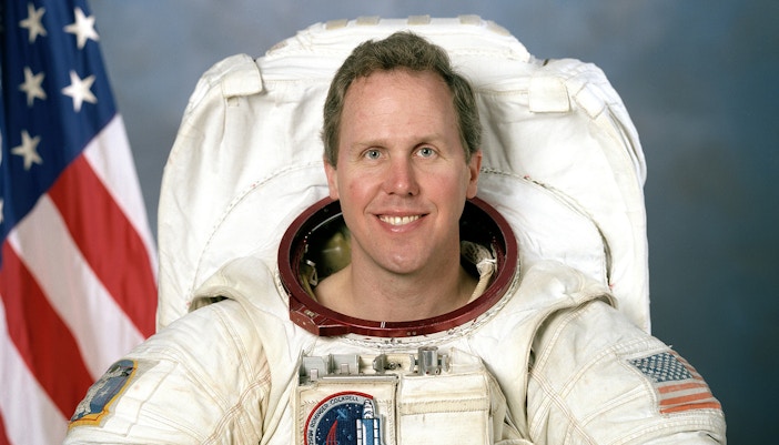 Astronaut Tom Jones in space suit with helmet, Earth visible in background.