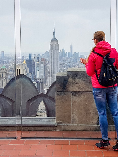 Woman with backpack viewing New York City skyline from rooftop, featuring Empire State Building.