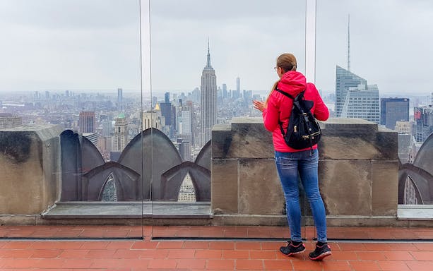 Woman with backpack viewing New York City skyline from rooftop, featuring Empire State Building.
