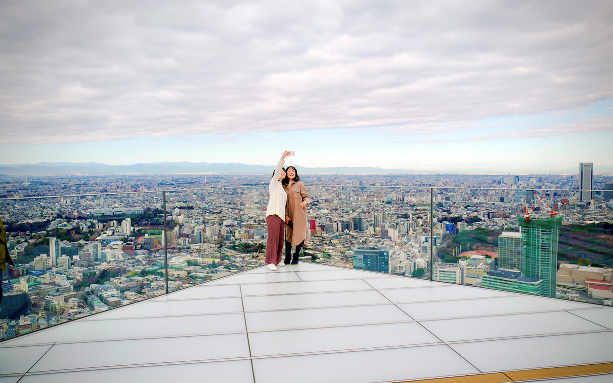 Tourists taking a selfie on Shibuya Sky Deck with Tokyo cityscape in the background.