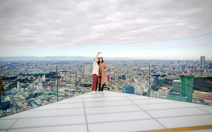 Tourists taking a selfie on Shibuya Sky Deck with Tokyo cityscape in the background.