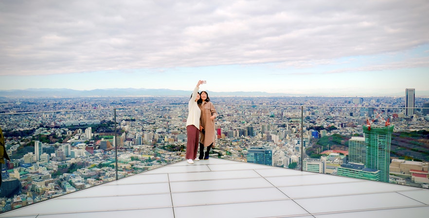 Tourists taking a selfie on Shibuya Sky Deck with Tokyo cityscape in the background.