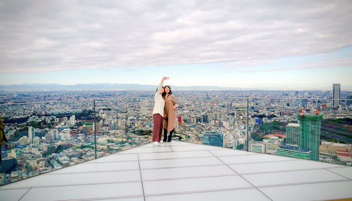 Tourists taking a selfie on Shibuya Sky Deck with Tokyo cityscape in the background.