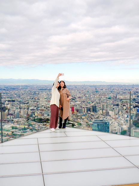 Tourists taking a selfie on Shibuya Sky Deck with Tokyo cityscape in the background.
