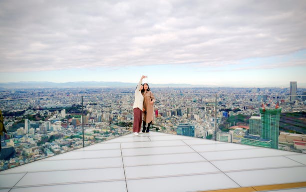 Tourists taking a selfie on Shibuya Sky Deck with Tokyo cityscape in the background.