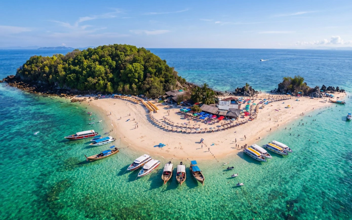 Speedboats docked at a sandy beach on Phi Phi Islands, Thailand, with tourists and colorful umbrellas.