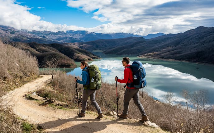 Hikers walking near Bovilla Lake with Gamti Mountains in the background.