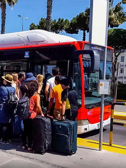 Passengers boarding a red bus with luggage at a sunny bus stop.